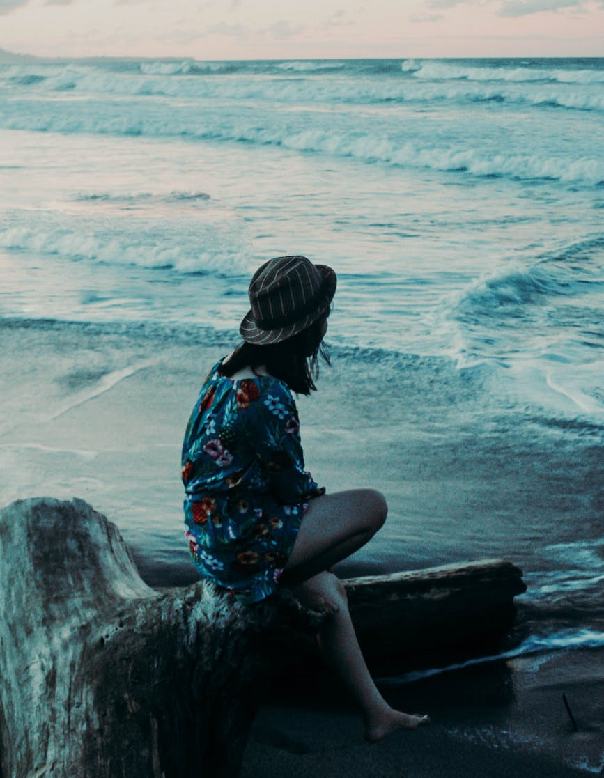 photo of woman in floral dress sitting alone on a log by the beach overlooking the horizon